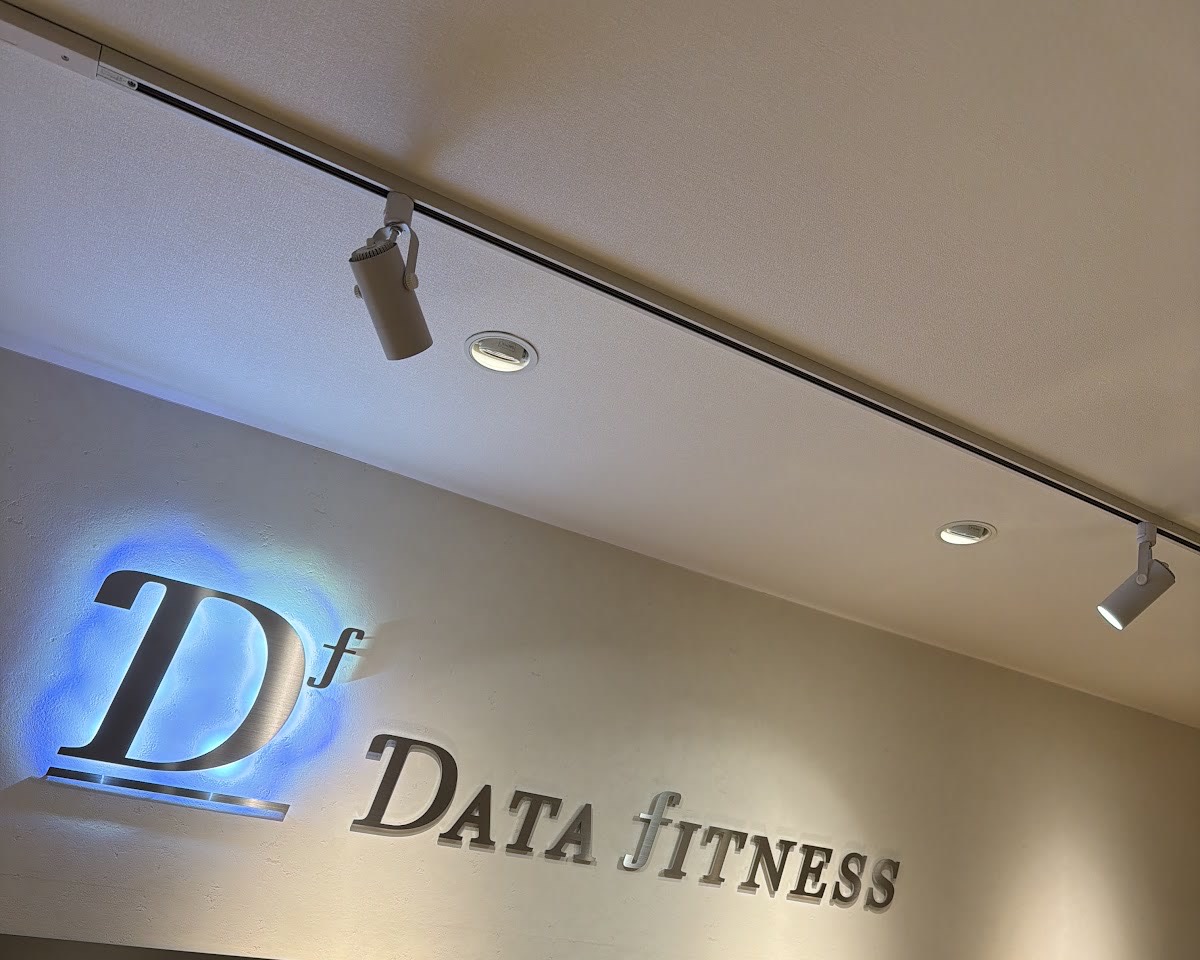 白い壁に取り付けられた、青く光る「Df」のモノグラムと「DATA fITNESS」の金属文字。スポットライトが照らす。