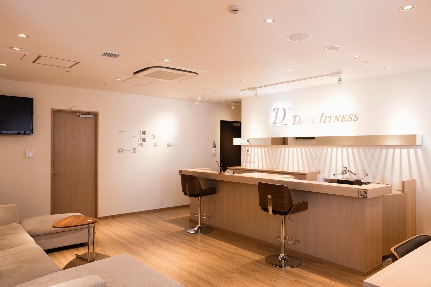 ベージュのソファと木目のカウンター、バースツール。奥の壁に「Df DATA fITNESS」のサイン。
